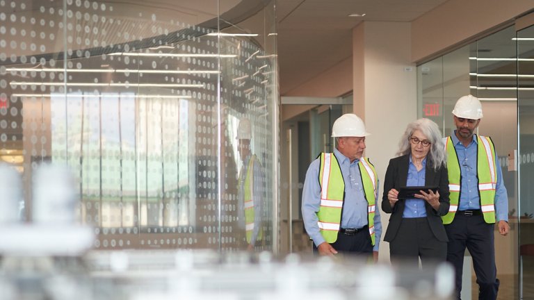 Two men in safety gear and one woman look at tablet in an office setting