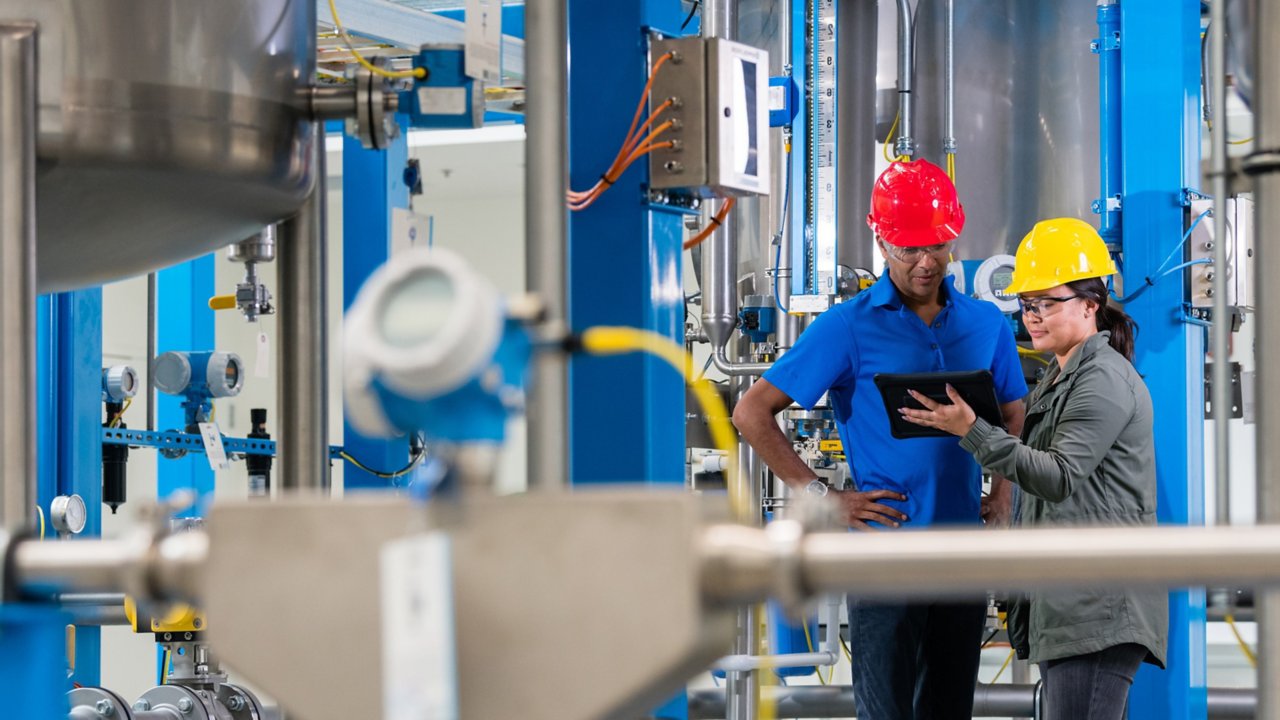 Two employees, a male and female wearing hard hats looking at a tablet in a plant