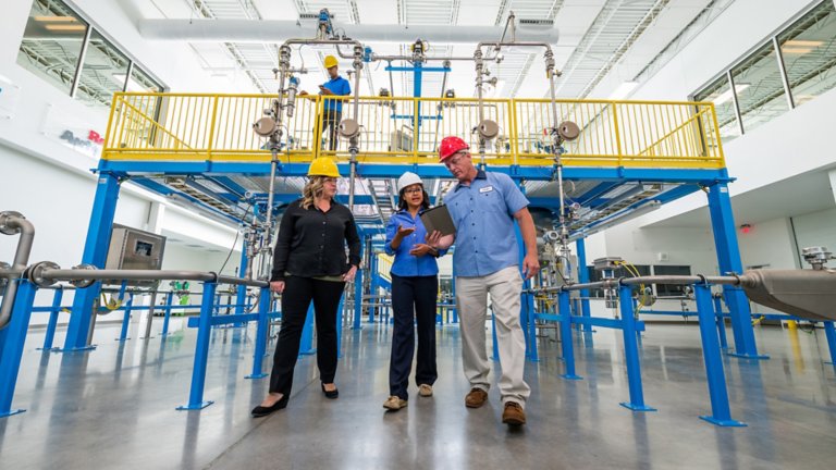 Two female employees and one male employee who is sharing information from a tablet he is holding while walking through an aisle in a factory.