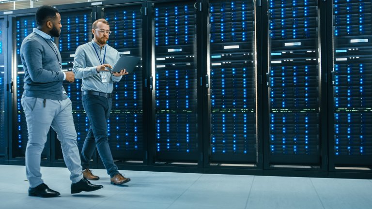  Two IT professionals walking through server room and consulting a laptop computer